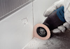 Gloved hand using an oscillating tool with a sanding attachment on a corner wall seam, dust scattered around.
