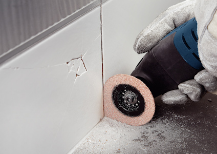 Gloved hand using an oscillating tool with a sanding attachment on a corner wall seam, dust scattered around.