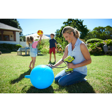 En person blåser upp en stor blå boll med en elverktygspump i en trädgård, medan två barn leker i bakgrunden.