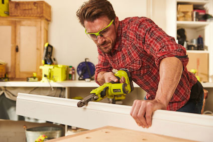 Person i rutig skjorta använder grön elverktyg för att såga genom en träregel i en verkstad.