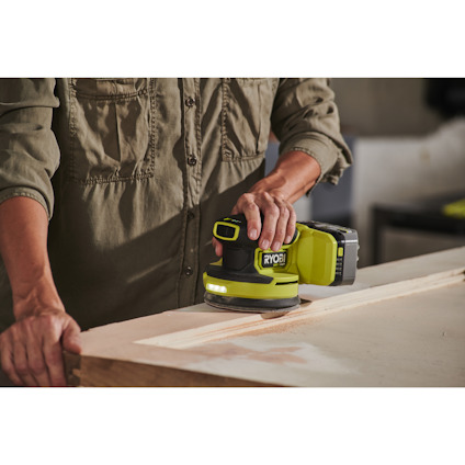 Person i skjorta använder en grön excenterslip från Ryobi på en träyta.