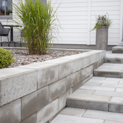 Gråa betongblock formar en låga mur intill stenlagda trappsteg, omgiven av växter och ett vitt hus.