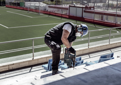 En person i skyddsutrustning bär ett gevär och går uppför läktartrappor med en tom fotbollsplan i bakgrunden.