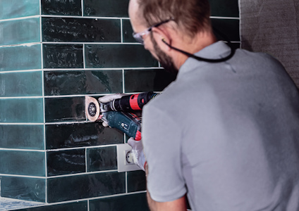 Person som använder borrmaskin på vägg med gröna kakelplattor.