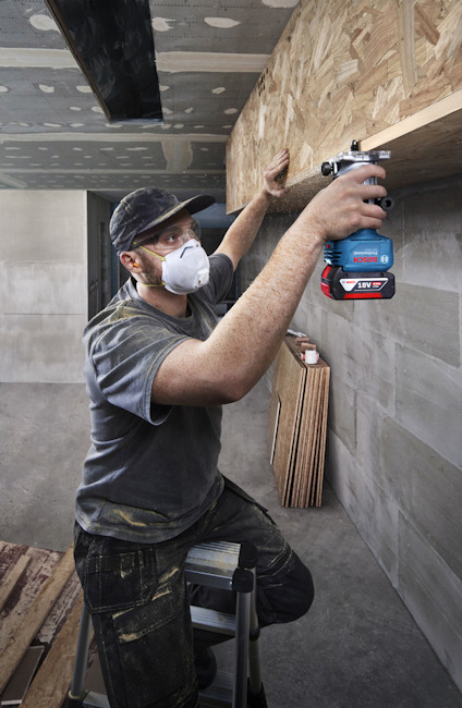 Person med ansiktsskydd och verktyg arbetar på en träpanel i ett rum under konstruktion.