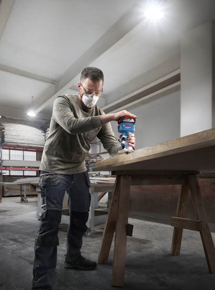 Person använder en handöverfräs på träsnitt i verkstad, iförd skyddsmask och arbetsbyxor.